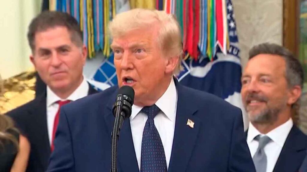 TRUMP-FOLARIN-BALOGUN-MUNDIAL - NL Directo Man in a navy suit speaks at a microphone, with two suited attendees and colorful ribbons behind him. The lapel pin resembles a flag badge.