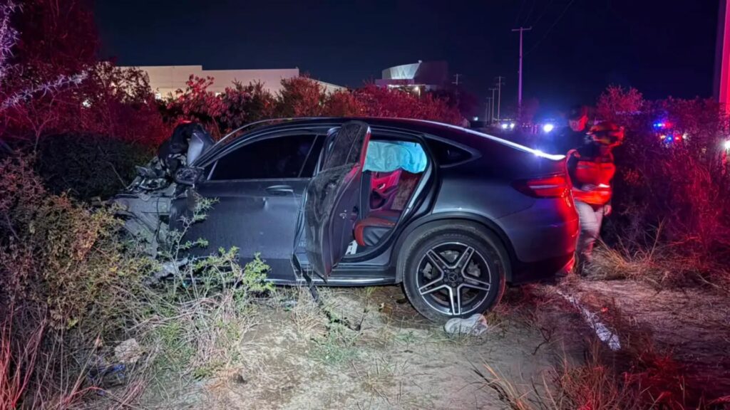 autopista-laredo-deja-un-muerto - NL Directo Nighttime car crash: a gray sedan with extensive front-end damage and an open rear-door, emergency responders in red gear nearby with flashing lights in the background.