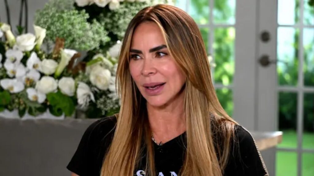 Woman with long, light brown hair speaking in an interview setting, floral arrangement behind her and glass doors visible in the background.