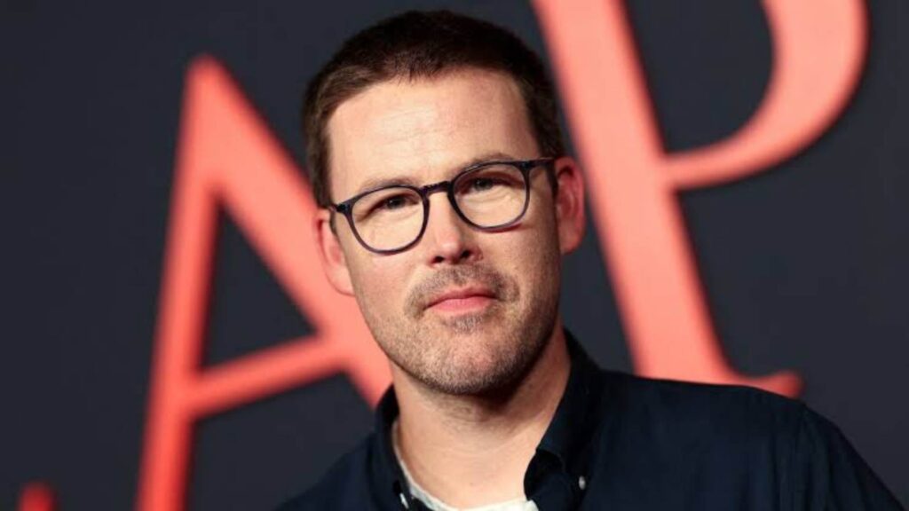 Portrait of a man with short dark hair and glasses, wearing a dark shirt, against a dark backdrop with orange/red geometric shapes behind him.