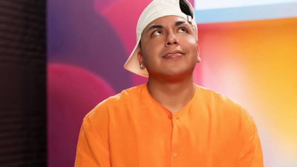 Person wearing an orange shirt and a white cap tilted to the side, looking upward with a slight smile against a colorful background.