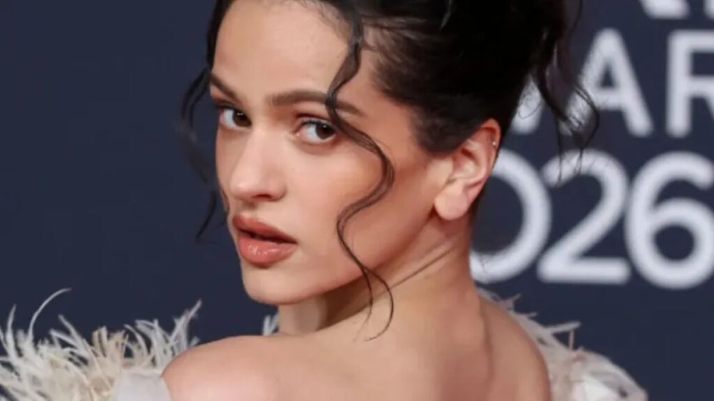 Close-up side profile of a woman with dark hair in an updo, wearing makeup and a feathered dress at a red-carpet event.
