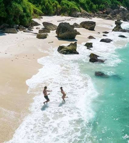 image - NL Directo Two people wading in shallow turquoise water at a sandy beach with rocks and jungle in the background.